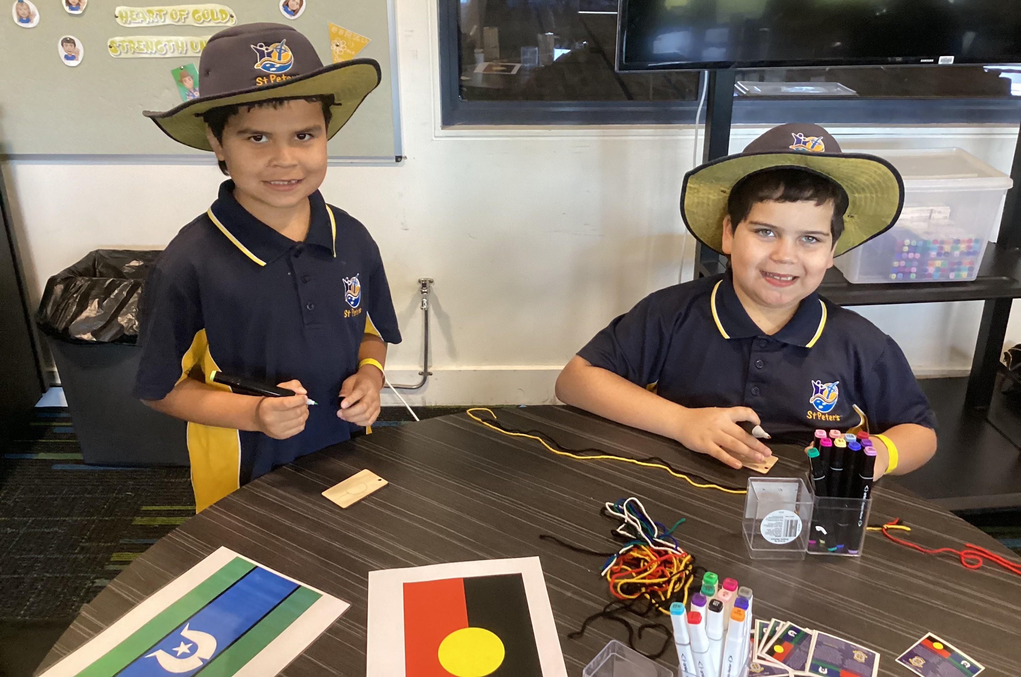 Two students in school uniforms sitting at a table with craft materials and printed Aboriginal and Torres Strait Islander flags