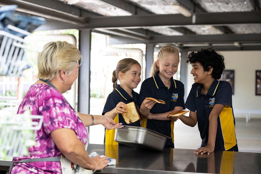 Students in school uniforms receiving food from an adult at an outdoor serving counter