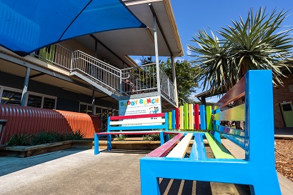 Outdoor area with a brightly painted bench near a school building and shade structure.