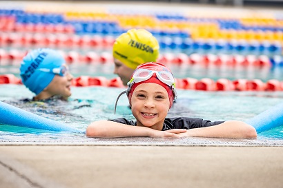 Child wearing a swim cap and goggles holding onto the pool edge, with other swimmers in the background